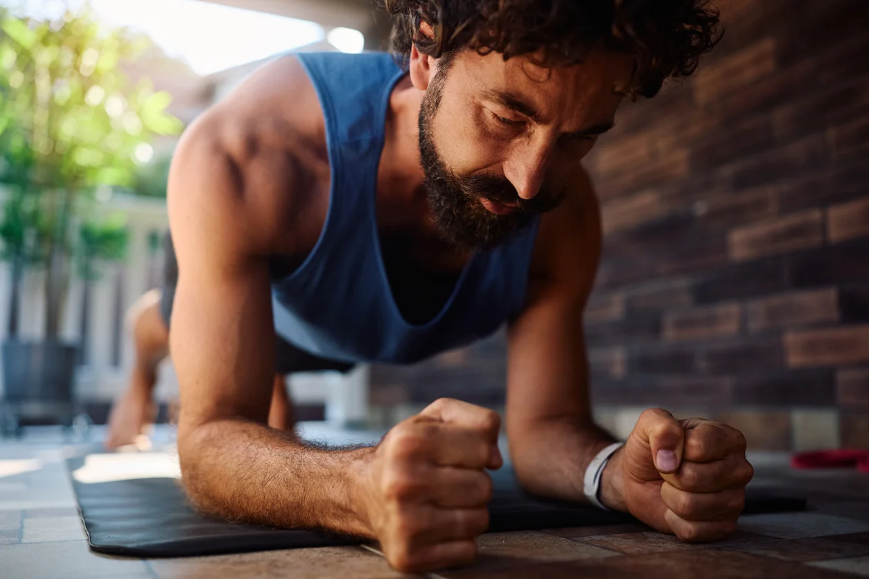 A man holding a forearm plank on a mat, eyes down, fists loose